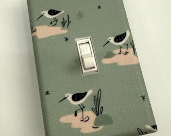 May include: A decorative light switch cover with a whimsical bird pattern. The cover is a muted green with illustrations of white and black birds, pink water, and black star accents. The switch is white and in the off position.