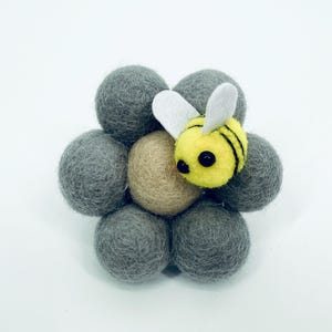 Pode incluir: Uma abelha de feltro feita à mão repousa sobre um grupo de bolas de feltro cinzentas, com uma bola bege no centro. A abelha é amarela com listras pretas e asas brancas. Perfeito para decoração de berçário ou como um presente único.