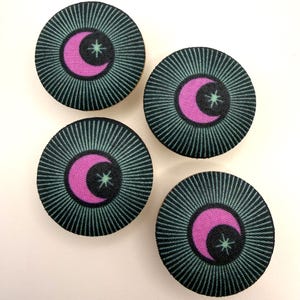 May include: Four circular fabric buttons with a celestial design. Each button features a pink crescent moon and star on a black background, surrounded by radiating teal lines. The buttons are arranged on a white surface.