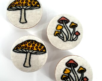May include: Four round fabric buttons with mushroom designs. The buttons feature yellow and red mushrooms with white stems on a white background.