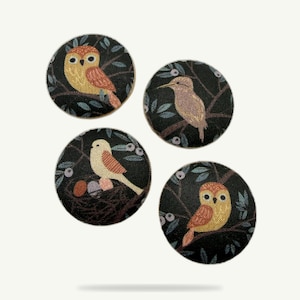 Op de afbeelding: Vier ronde stoffen knopen met vogelontwerpen. Elke knoop heeft een andere vogelillustratie in tinten geel, oranje en bruin tegen een donkere achtergrond met bladeren en bessen. De knopen hebben een diameter van ongeveer 2,5 cm.