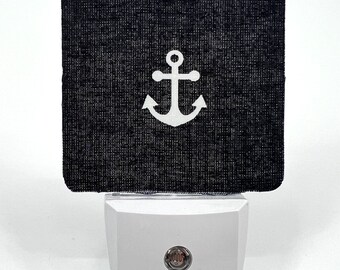 May include: A white anchor on a black fabric nightlight plug-in. The nightlight is plugged into a standard electrical outlet.