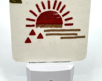 May include: A white night light with a beige square insert featuring a red and brown geometric sun design. The sun has a red center and brown rays. There are three red triangles at the bottom of the design.