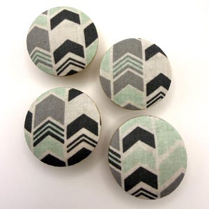 Op de afbeelding: Vier ronde, met stof beklede knopen met een geometrisch pijlenpatroon in grijstinten, zwart en mintgroen. De knopen zijn gerangschikt op een witte ondergrond, waarbij hun decoratieve ontwerp wordt getoond.