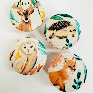 Könnte beinhalten: Vier runde Stoffknöpfe mit weißem Hintergrund und einem Waldtier-Design. Die Knöpfe zeigen ein Reh, einen Igel, eine Eule und einen Fuchs.