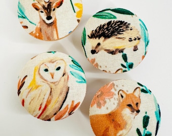 May include: Four round fabric buttons with a white background and a woodland animal design. The buttons feature a deer, a hedgehog, an owl, and a fox.
