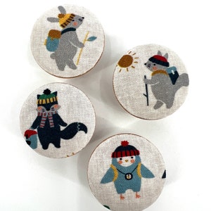 May include: Four round fabric buttons with different animal designs. Each button features a different animal wearing a hat and carrying a backpack. The animals include a rabbit, a squirrel, a fox, and a penguin.