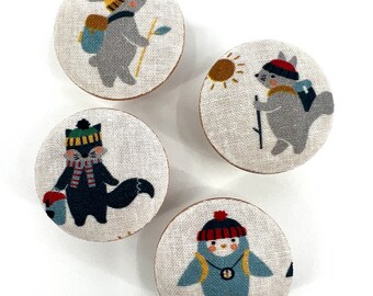 May include: Four round fabric buttons with different animal designs. Each button features a different animal wearing a hat and carrying a backpack. The animals include a rabbit, a squirrel, a fox, and a penguin.