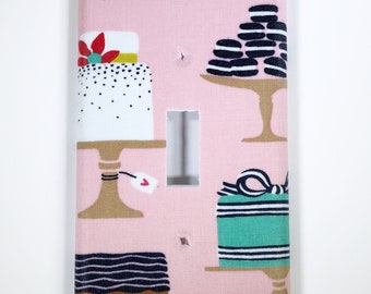 May include: A pink light switch cover with a pattern of cakes, cookies, and presents. The cakes are decorated with white frosting and black sprinkles. The cookies are black and white. The presents are wrapped in green paper with white stripes and a black bow.