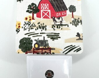 May include: A white night light with a red barn, green trees, and a cow on a beige background. The night light has a small, round, silver button on the front.