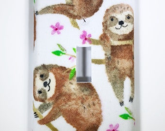 May include: A white light switch cover featuring a repeating pattern of cartoon sloths hanging from branches. The sloths are brown with tan accents, and the branches and leaves are green. Pink flowers are scattered across the background.