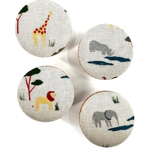 Puede incluir: Cuatro pomos redondos de tela con diferentes diseños de animales. Los pomos presentan una jirafa, un león, un rinoceronte y un elefante. Los pomos son beige con acentos verdes, rojos y amarillos.