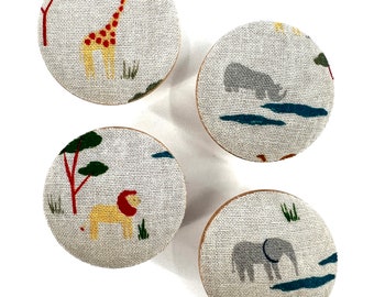 May include: Four round fabric knobs with different animal designs. The knobs feature a giraffe, a lion, a rhino, and an elephant. The knobs are beige with green, red, and yellow accents.