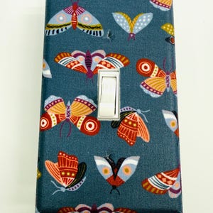May include: A decorative light switch cover featuring a pattern of colorful moths on a teal background. The moths are in shades of red, orange, yellow, and blue. The switch itself is white.