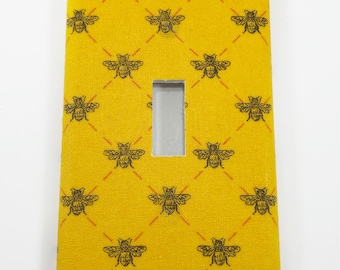 May include: A yellow light switch cover with a repeating pattern of black bees. The switch cover has a rectangular cutout for the light switch.