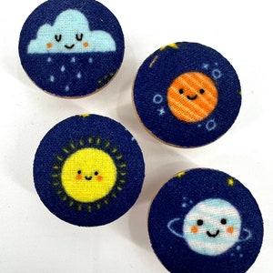 Puede incluir: Cuatro botones redondos de tela con fondo azul y diferentes diseños con temática espacial. Los diseños incluyen una nube con gotas de lluvia, un sol, un planeta con anillos y un planeta rayado.