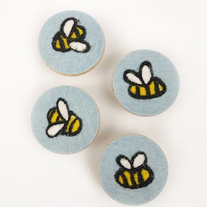 Könnte beinhalten: Vier runde blaue Stoffornamente mit gestickten gelben und schwarzen Bienen. Jedes Ornament hat ein anderes Bienendesign.