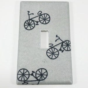 Könnte beinhalten: Ein hellgrauer Lichtschalter mit einem schwarz-weißen Fahrrad-Design. Das Design wird dreimal auf dem Schalter wiederholt.