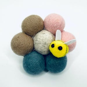 Pode incluir: Um conjunto de bolas de feltro em tons de castanho, rosa, azul-petróleo e creme, dispostas em torno de uma abelha de feltro amarela com olhos pretos e asas brancas. A abelha é um acréscimo divertido.