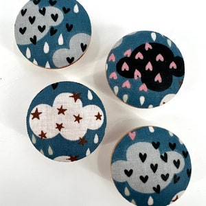 Puede incluir: Cuatro pomos redondos de madera con diferentes diseños de tela que presentan nubes, estrellas y corazones. Los pomos son azules con nubes blancas y acentos de varios colores.