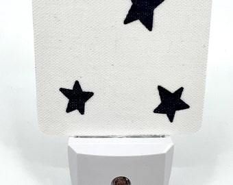 May include: White night light with a black fabric shade featuring three black stars. The night light has a white plastic base with a small red light.