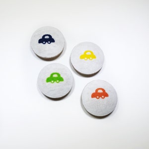 Puede incluir: Cuatro botones redondos de tela con fondo blanco y un pequeño diseño de coche en diferentes colores: azul, amarillo, verde y naranja.