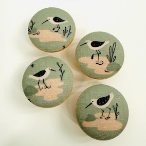 May include: Four round, fabric-covered buttons with a nature theme. Each button features a stylized illustration of a bird with black and white markings standing on a tan-colored patch, set against a muted green background.