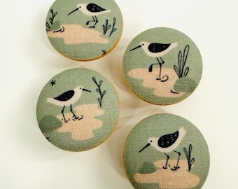 May include: Four round, fabric-covered buttons with a nature theme. Each button features a stylized illustration of a bird with black and white markings standing on a tan-colored patch, set against a muted green background.