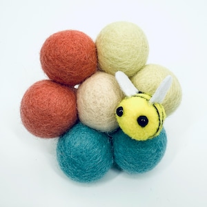 Pode incluir: Uma coleção de bolas de feltro coloridas em tons de laranja, azul-petróleo, creme e amarelo. Uma pequena abelha de feltro amarelo com listras pretas e asas brancas está aninhada entre as bolas. O arranjo está em um fundo branco.