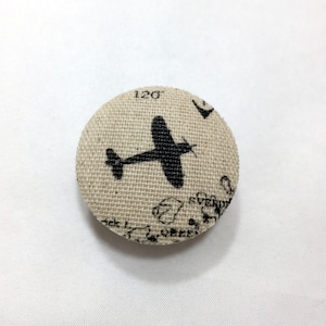 May include: A round, beige fabric button with a black silhouette of an airplane printed on it. The button has a textured surface and a white background with black text.