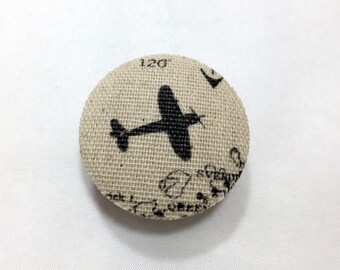 May include: A round, beige fabric button with a black silhouette of an airplane printed on it. The button has a textured surface and a white background with black text.