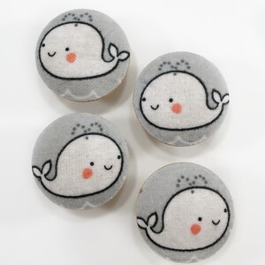 May include: Four grey and white fabric buttons featuring a whale design. Each button depicts a white whale with a black outline and a small red dot on its cheek. The whale is smiling and has a small spout on its head. These buttons are ideal for adding a touch of whimsy to any project.