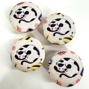 May include: Four round wooden knobs with a white fabric covering featuring a black and white cat design. The fabric has a colourful geometric pattern with pink, yellow, and blue accents.