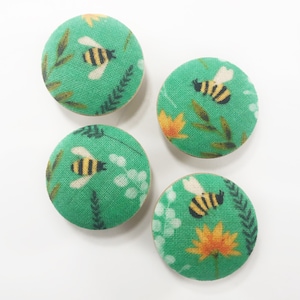 Może przedstawiać: Cztery zielone guziki z tkaniny z wzorem pszczół i kwiatów. Guziki są okrągłe i mają białe tło z zielonymi liśćmi i żółtymi kwiatami. Pszczoły są czarne i żółte.