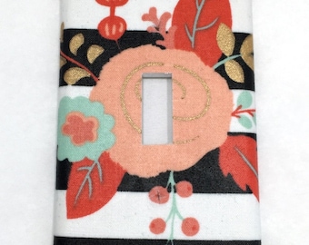 May include: A decorative light switch cover featuring a floral design. The cover has a black and white striped background with a large peach-colored flower and red leaves. Gold accents and a mint green flower add detail.
