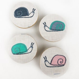May include: Four round fabric ornaments with a white background and a black outline of a snail with different colored shells. The shells are pink, green, gray, and black.
