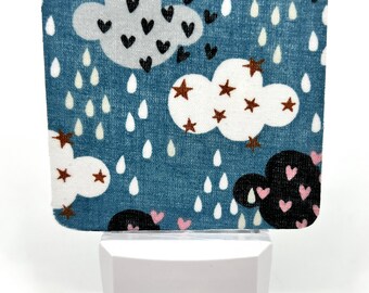 May include: A white night light with a blue and white fabric cover featuring a pattern of clouds, stars, and raindrops. The fabric cover has pink and black hearts.