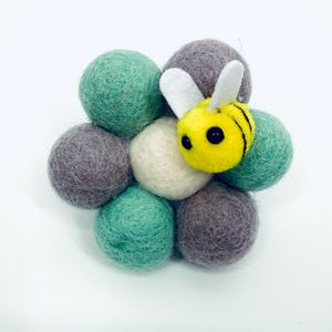 Pode incluir: Um conjunto de bolas de feltro em tons de verde, cinza e creme, com uma abelha amarela e preta presa. A abelha tem asas brancas e olhos pretos. As bolas de feltro são dispostas em um padrão floral sobre um fundo branco.