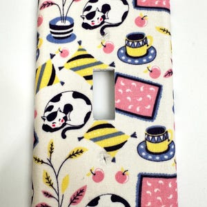 May include: A white light switch cover with a colorful pattern of cats, plants, cups, and geometric shapes. The pattern is in shades of pink, yellow, blue, and black.