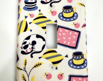May include: A white light switch cover with a colorful pattern of cats, plants, cups, and geometric shapes. The pattern is in shades of pink, yellow, blue, and black.
