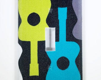 May include: A decorative light switch cover featuring a repeating pattern of stylized guitars in shades of green, blue, and gray on a black background. The cover has a rectangular shape with a cutout for the switch.