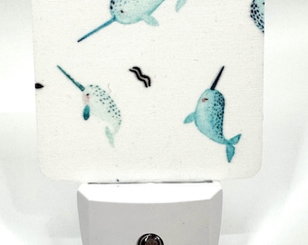 May include: A white night light featuring a repeating pattern of blue narwhals. The night light has a square, fabric-covered face with the narwhal design and a white plastic base with a small, circular light sensor.