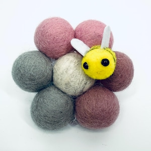 Pode incluir: Uma abelha de feltro artesanal repousa sobre um grupo de bolas de feltro. As bolas são em tons de rosa, cinza e marrom. A abelha é amarela com listras e olhos pretos, e asas brancas.