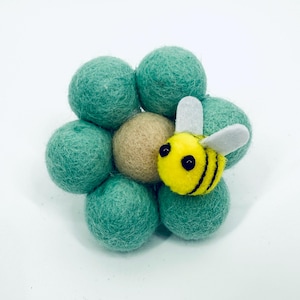 Pode incluir: Uma abelha de feltro feita à mão repousa sobre um grupo de bolas de feltro verdes, com uma bola bege no centro. A abelha é amarela com listras pretas e asas brancas. As bolas de feltro estão dispostas em um padrão circular.