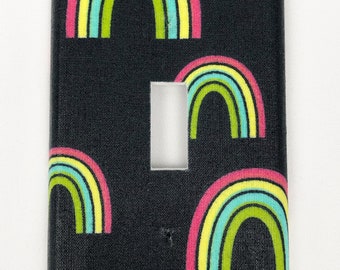May include: A black light switch cover with a repeating pattern of colorful rainbows. The rainbows are pink, yellow, green, and blue.