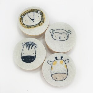 Puede incluir: Cuatro posavasos redondos de tela blanca con diferentes diseños de animales. Los posavasos presentan un león, un mono, una cebra y una jirafa.