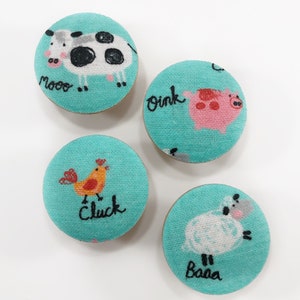 Puede incluir: Cuatro botones redondos de tela con diferentes diseños de animales de granja. Los botones son de color azul turquesa con costuras blancas. Los diseños incluyen una vaca con la palabra "moo", un cerdo con la palabra "oink", una gallina con la palabra "cluck" y una oveja con la palabra "baa".