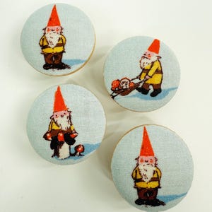 May include: Four round buttons with light blue fabric and wooden backs. Each button features a cartoon gnome with a red hat, yellow jacket, and brown pants. One gnome is pushing a wheelbarrow.