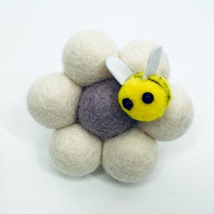 May include: A handmade felt bee rests on a cluster of cream-colored felt balls, with a central purple ball. The bee is yellow with black eyes and white wings. This whimsical piece is perfect for home decor or as a unique gift.