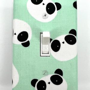 Op de afbeelding: Een lichtschakelaar-afdekplaat met een groene achtergrond en een patroon van zwart-witte panda gezichten. De schakelaar is wit en heeft de tekst "ON".